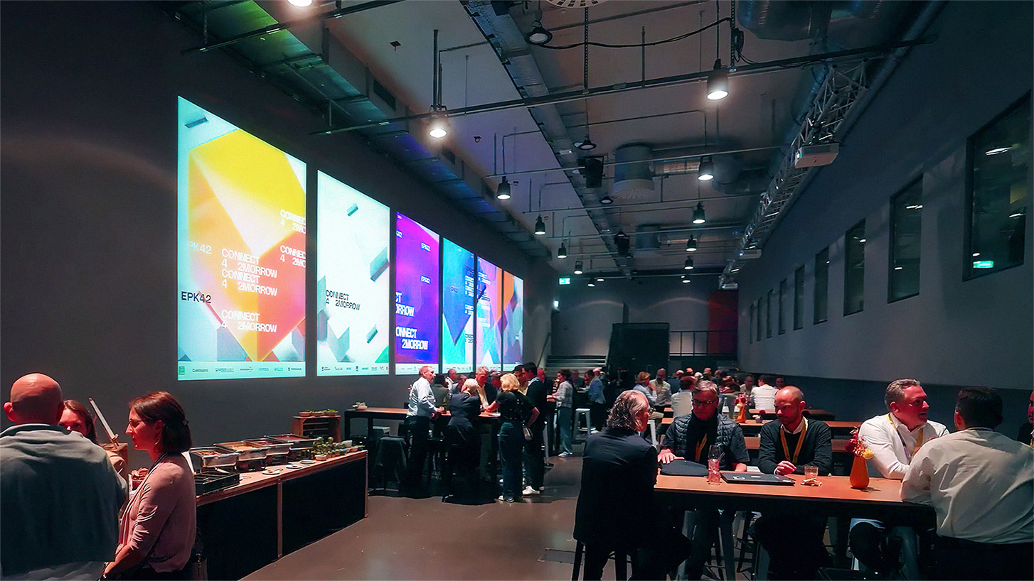 Abendliches Networking-Event in einer modernen Halle mit hoher Decke und industriellem Charakter. An der linken Wand sind großformatige Projektionen mit dem farbenfrohen Eventdesign „EPK42 – CONNECT 4 2MORROW“ zu sehen. Zahlreiche Gäste sitzen oder stehen an hohen Tischen, unterhalten sich und genießen Speisen vom Buffet. Die Stimmung ist lebendig und professionell.