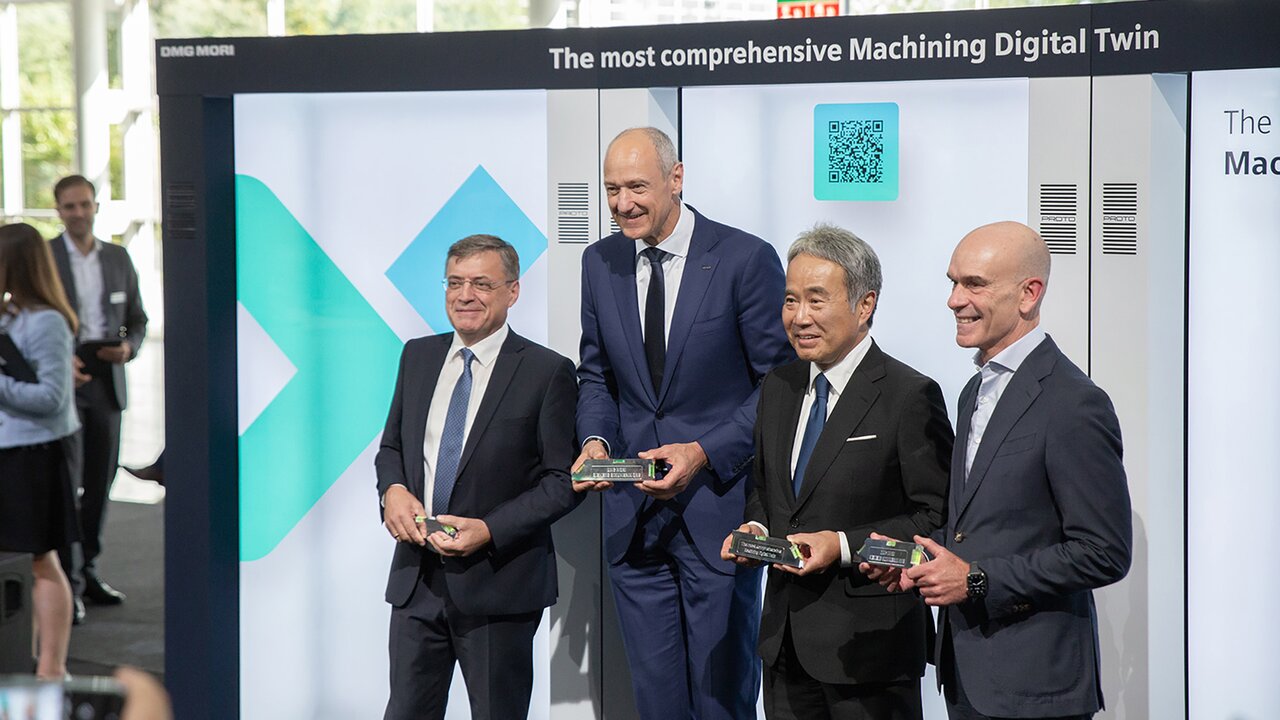 Vier Männer im Anzug stehen nebeneinander vor einer Messewand mit dem Schriftzug ‚The most comprehensive Machining Digital Twin‘.