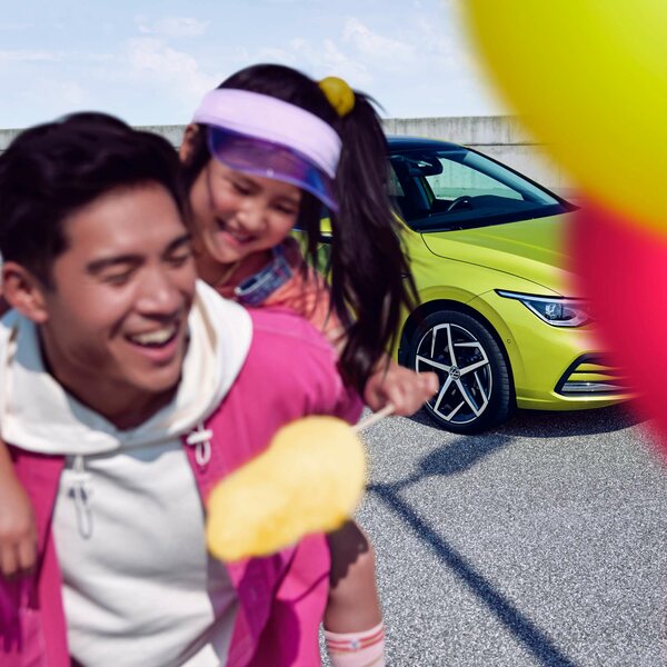 Mann trägt lachendes Mädchen auf dem Rücken, im Hintergrund ein gelber VW.