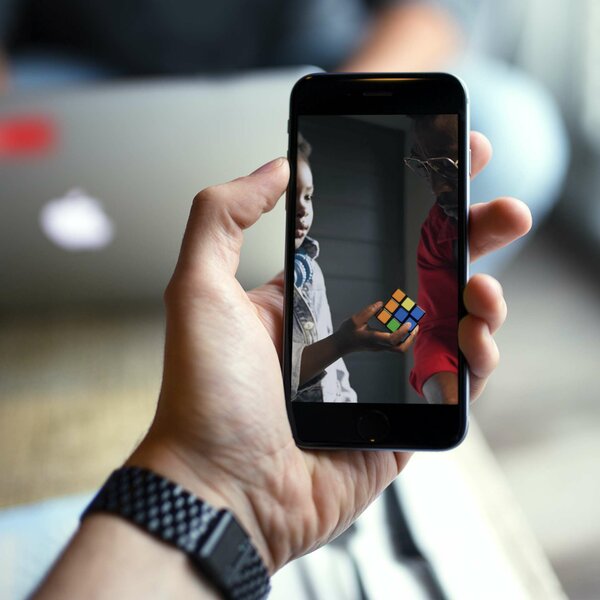 Hand hält Smartphone mit Bild eines Kindes und Erwachsenen, die gemeinsam einen Zauberwürfel betrachten.