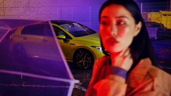 Frau unter transparentem Regenschirm, im Hintergrund ein gelber VW bei Nacht.