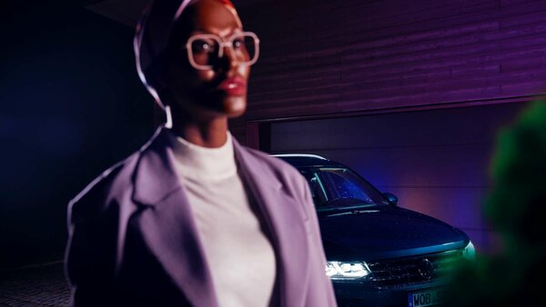 Frau mit Brille im Vordergrund, dahinter ein VW mit eingeschaltetem Licht bei Nacht.