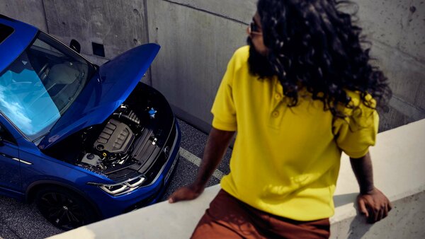 Mann mit langen Haaren blickt auf ein Auto mit offenem Motorraum in einem Parkdeck.