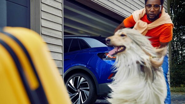Mann mit Hund vor einer geöffneten Garage, in der ein blauer VW steht; gelber Koffer im Vordergrund.