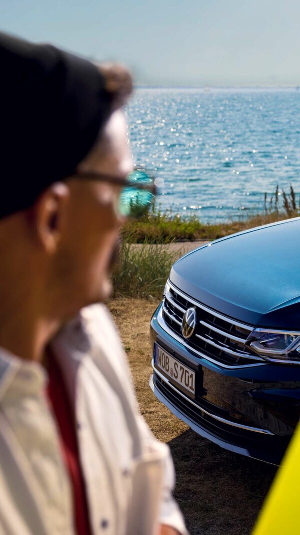 Mann mit Sonnenbrille blickt aufs Meer, im Hintergrund ein schwarzer VW, auf dessen Motorhaube sich der blaue Himmel spiegelt.