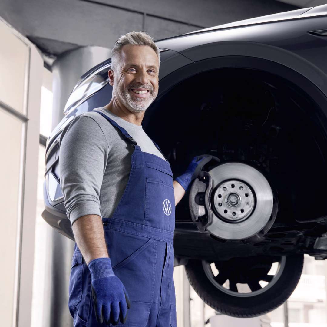 Lächelnder Mann in blauer VW-Arbeitskleidung steht neben einem aufgebockten Auto und zeigt auf die Bremsscheibe. Die Szene spielt in einer hellen Werkstatt.