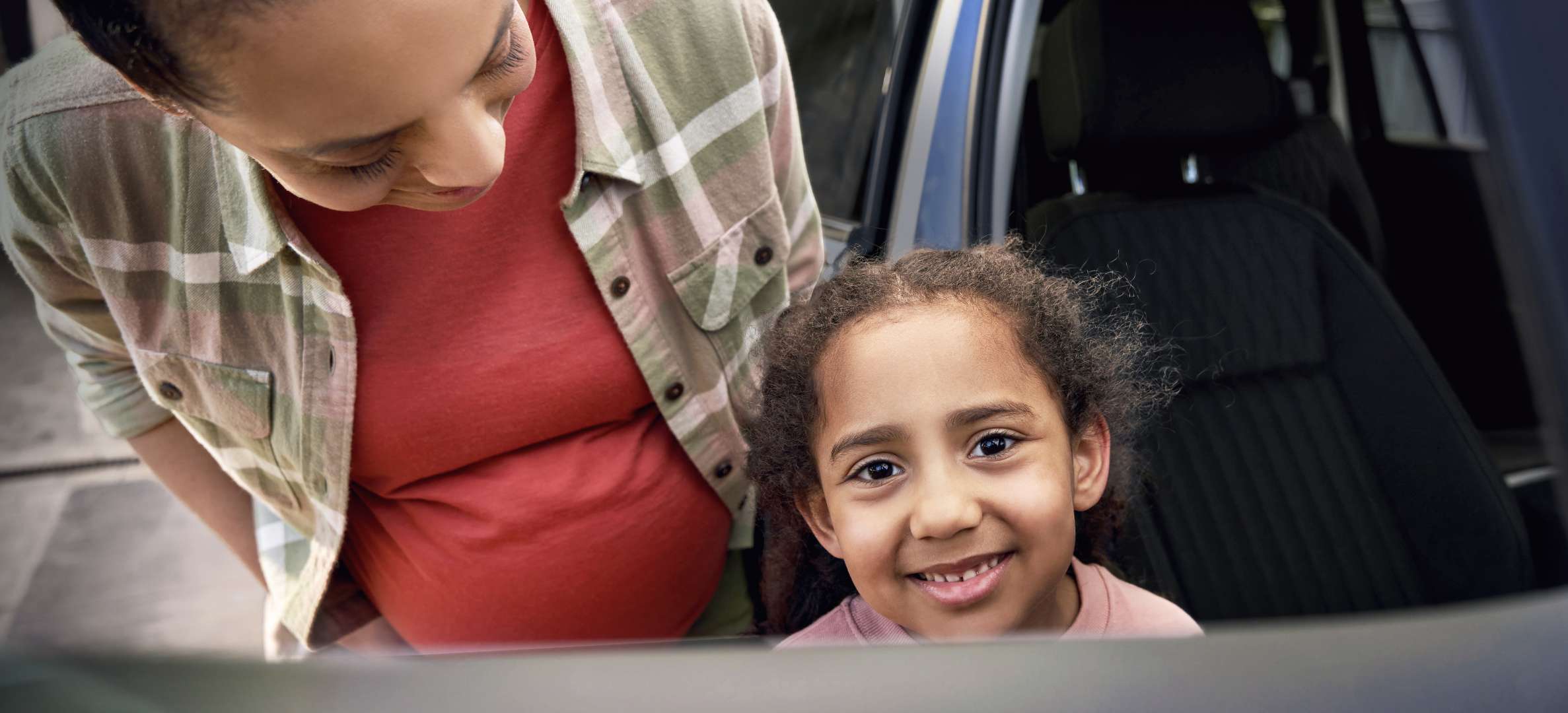 Nahaufnahme einer Mutter, die sich liebevoll über ihre Tochter beugt. Das Mädchen sitzt im Auto, lächelt direkt in die Kamera. Beide wirken fröhlich und vertraut.
