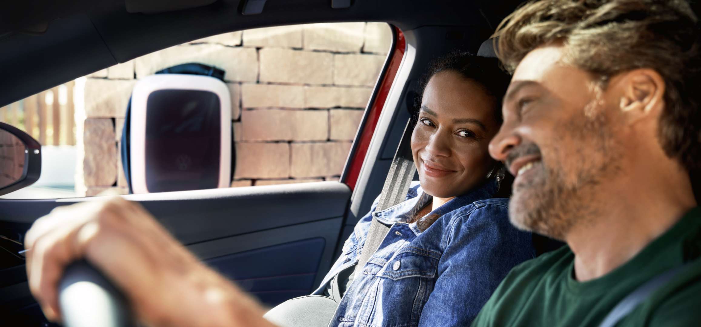 Paar sitzt im Auto, die Frau schaut lächelnd zum Fahrer. Im Hintergrund sieht man eine VW-Wallbox an einer Steinwand – Hinweis auf E-Mobilität. Die Szene vermittelt Nähe und Alltag.