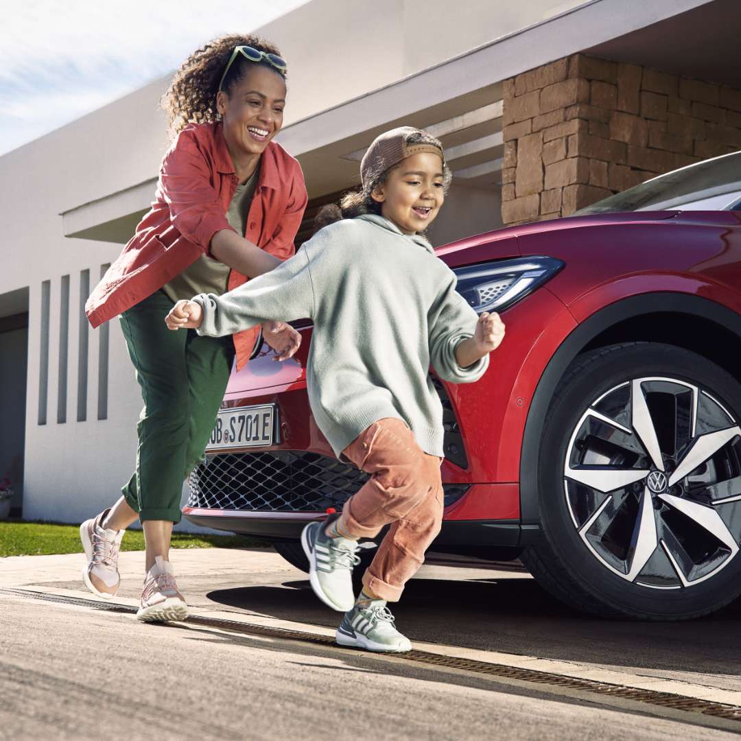 Fröhliche Szene vor einem roten VW-Elektroauto: Eine Frau läuft lachend ihrem Kind hinterher, das ausgelassen in Richtung Kamera rennt. Im Hintergrund ein modernes Haus mit Garage.