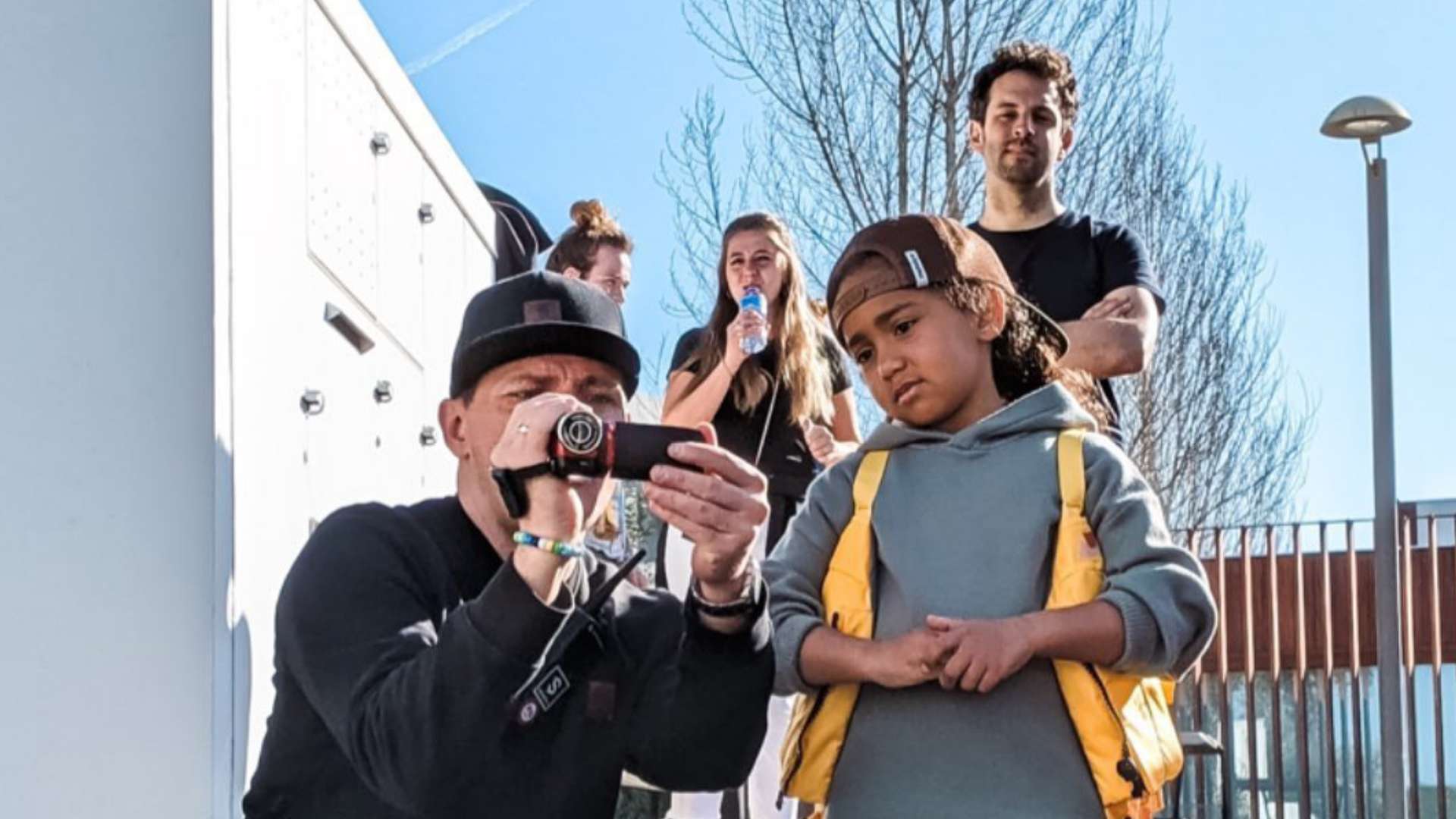 Ein Kameramann im schwarzen Outfit zeigt einem Kind mit gelber Weste und Basecap eine Videoaufnahme auf einem Camcorder. Im Hintergrund stehen drei weitere Personen vor einem modernen Gebäude und beobachten die Szene.