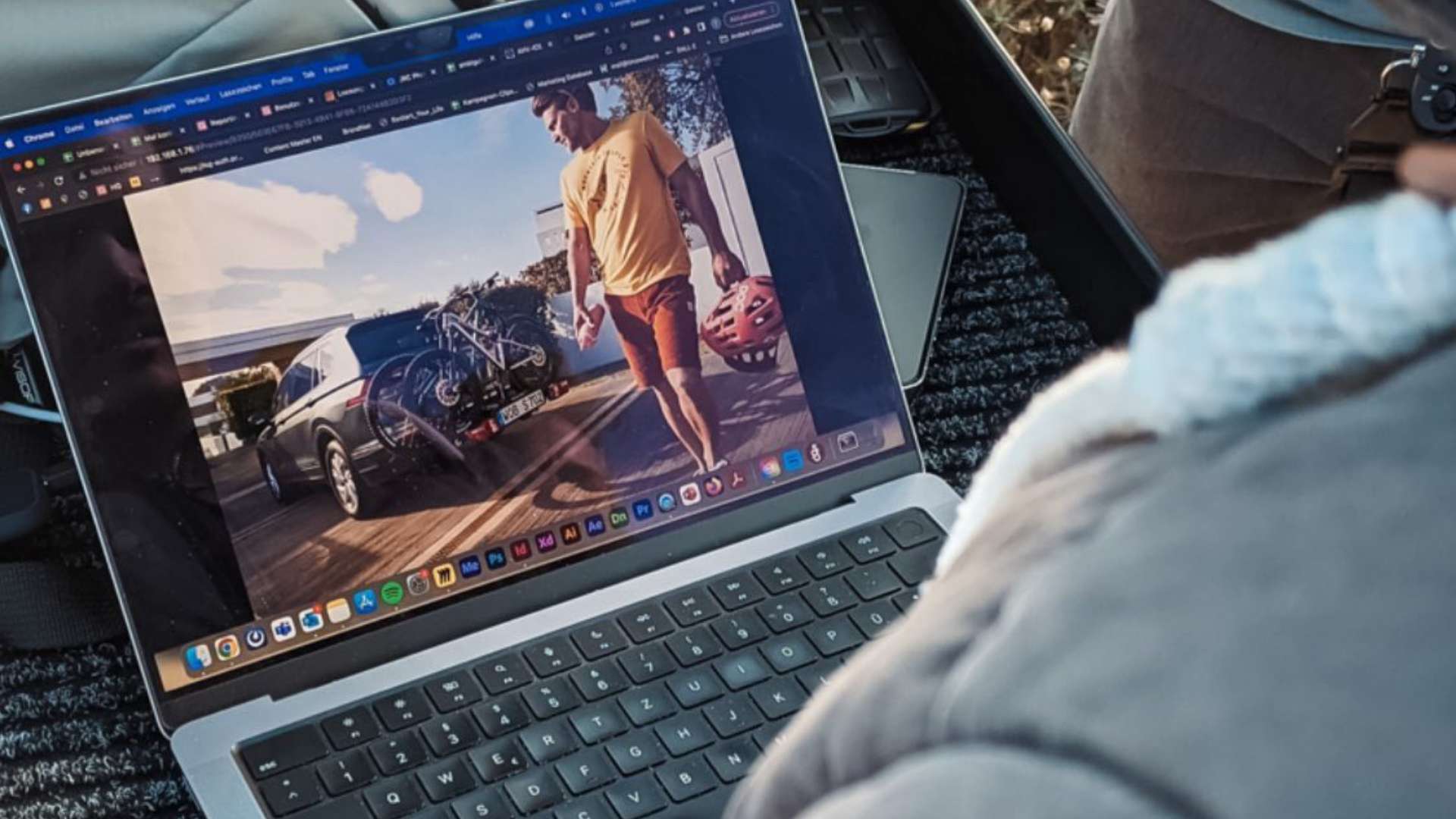 Detailaufnahme eines Laptops mit geöffneter Bildbearbeitungssoftware. Auf dem Bildschirm ist ein Bild zu sehen, das einen Mann mit Fahrradhelm neben einem Auto mit Fahrradträger zeigt. Der Laptop steht auf einem dunklen Teppichboden im Kofferraum.
