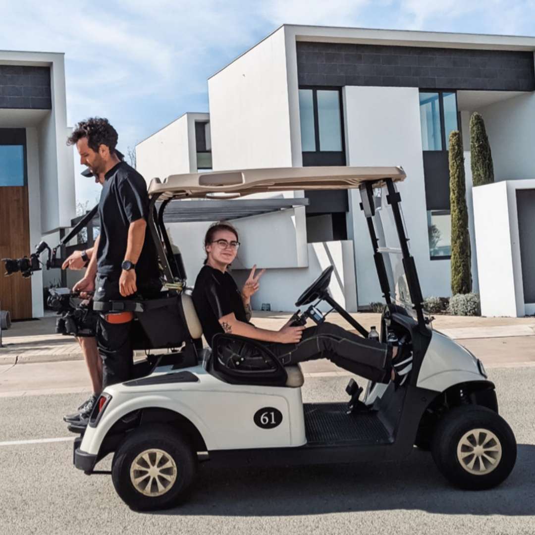 Zwei Personen vom Filmteam fahren mit einem Golfcart durch eine Wohnsiedlung mit moderner Architektur. Eine Person filmt im Stehen auf der Rückbank, die andere fährt und zeigt ein Peace-Zeichen. Beide tragen schwarze Kleidung.