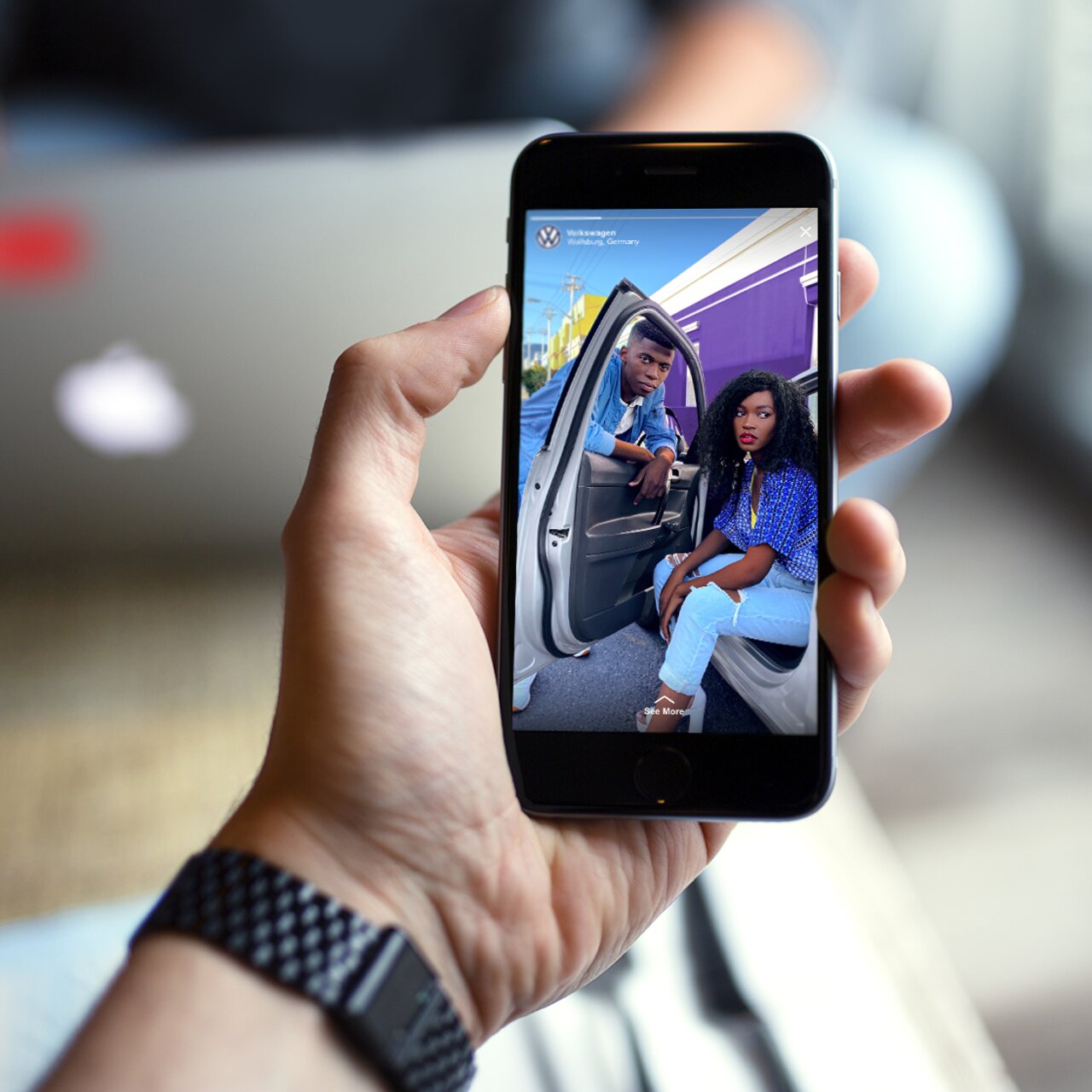 Hand hält Smartphone mit geöffnetem Instagram-Post von zwei Personen am Auto vor bunter Hausfassade.