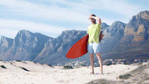 Mann mit Surfbrett steht barfuß am Strand und blickt in die Ferne; dahinter hohe Bergkette.