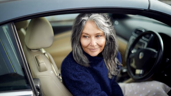 Grauhaarige Frau mit Pullover sitzt lächelnd auf dem Fahrersitz eines Autos mit beigem Innenraum.