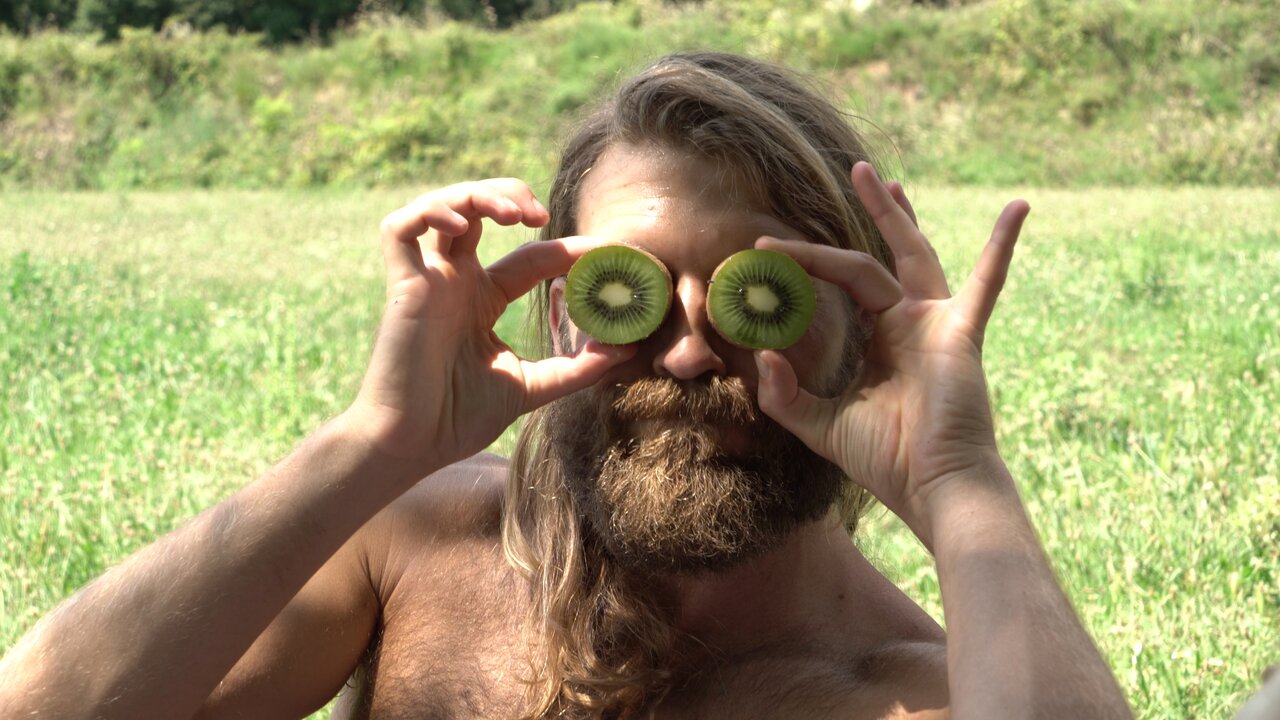 Mann mit langem Haar hält zwei Kiwi-Hälften vor seine Augen und lächelt dabei verspielt.