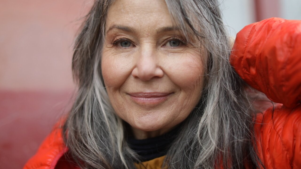 Ältere Frau mit grauen Haaren und roter Jacke lächelt ruhig in die Kamera.