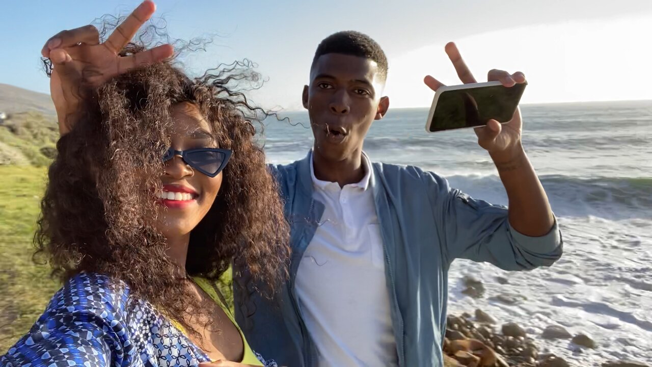 Zwei junge Erwachsene machen Selfie am Meer, zeigen Peace-Zeichen und lächeln fröhlich.