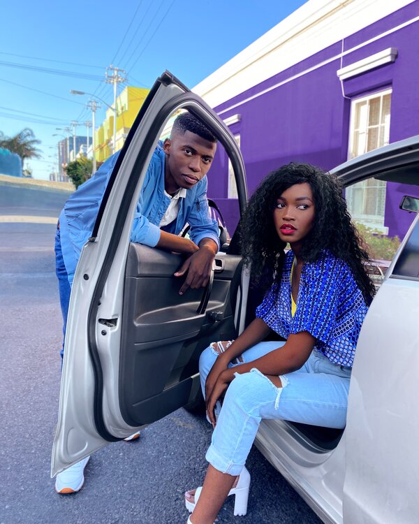 Zwei junge Erwachsene posieren am offenen Autotürrahmen vor einer farbigen Hausfassade.