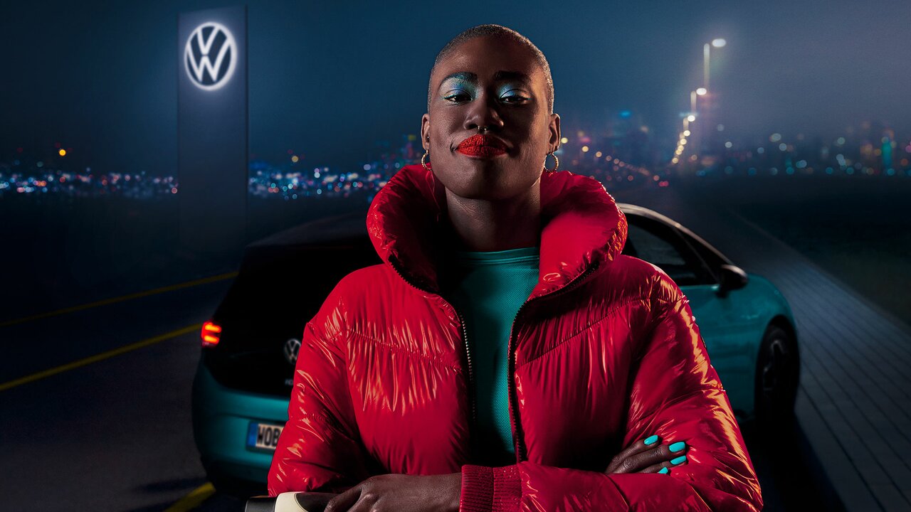 Selbstbewusste Frau in roter Jacke steht vor einem VW bei Nacht mit beleuchtetem VW-Logo im Hintergrund.