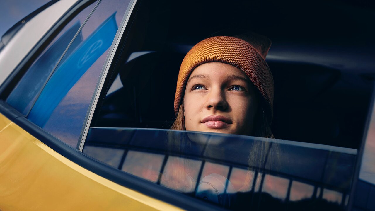 Mädchen mit Mütze schaut aus dem Fenster eines gelben Autos, im Hintergrund spiegeln sich Gebäude und Himmel.