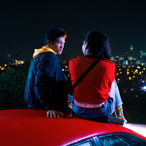 Blick aus einem Auto auf einen Mann mit Brille, der nachdenklich an einem türkisfarbenen See mit bewaldeten Hügeln steht.