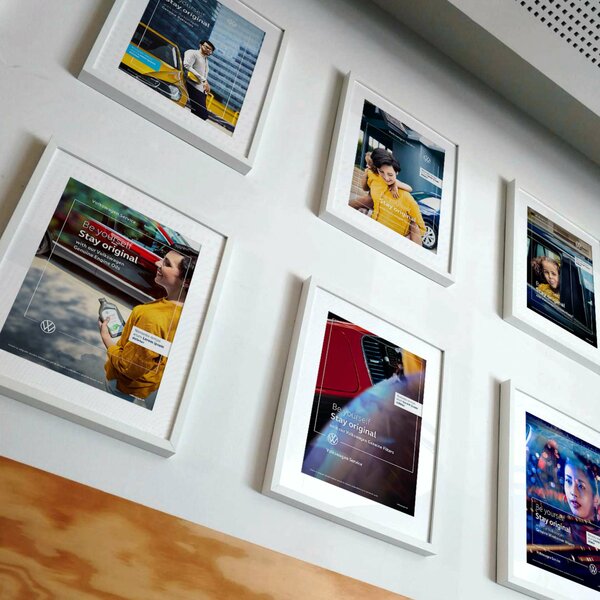 Sechs gerahmte Volkswagen-Poster hängen an einer Wand, alle zeigen unterschiedliche Personen mit Autos und dem Slogan „Be yourself. Stay original.“