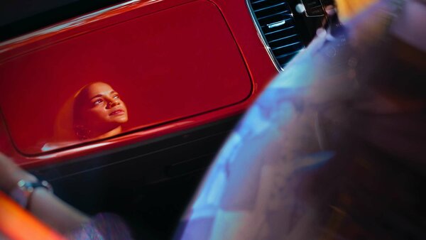 Das Gesicht einer Frau spiegelt sich auf der glänzenden roten Armaturenbrett-Verkleidung eines Autos wider; der Blick geht verträumt nach oben.