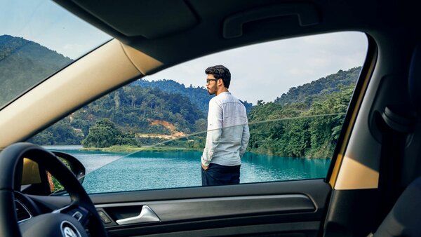 Frau mit Kind auf dem Rücken lacht; sie trägt ein senfgelbes Hemd und steht vor einem dunkelblauen SUV.