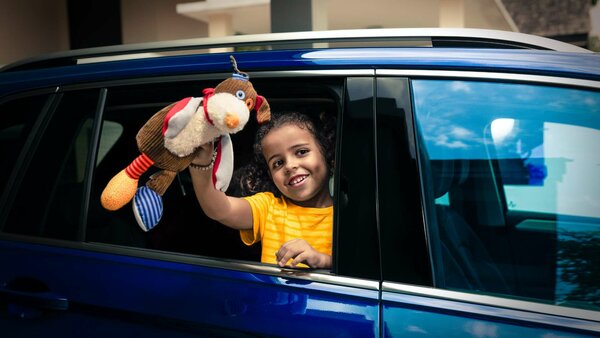 Mädchen im gelben T-Shirt schaut lachend aus dem geöffneten Autofenster eines blauen Fahrzeugs und hält ein buntes Stofftier in die Höhe.