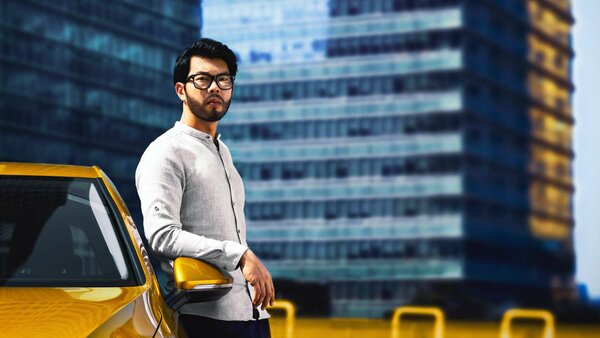 Ein Mann mit Brille steht selbstbewusst an einem gelben Auto vor einem modernen Bürogebäude.