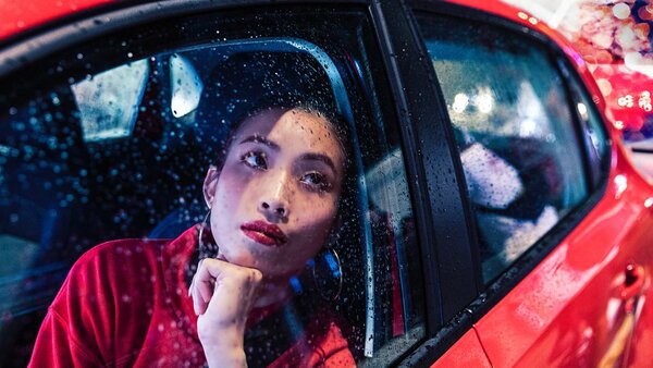 Eine junge Frau sitzt nachdenklich bei Regen auf dem Rücksitz eines roten Autos und schaut aus dem Fenster.