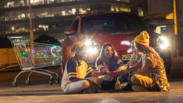 Drei junge Frauen sitzen lachend bei Nacht vor einem Auto, neben ihnen ein beleuchteter Einkaufswagen.