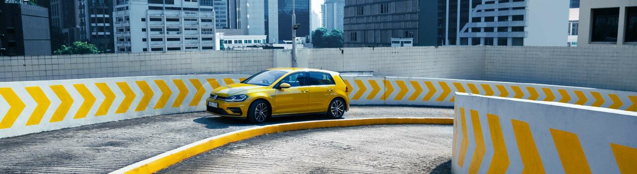 Gelber Kompaktwagen fährt auf einer spiralförmigen Parkhausrampe inmitten einer Hochhauskulisse mit gelben Richtungspfeilen an der Mauer.