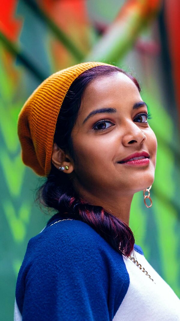 Junge Frau mit orangefarbener Mütze und großen Ohrringen blickt selbstbewusst zur Kamera; im Hintergrund eine bunt verschwommene Wand.