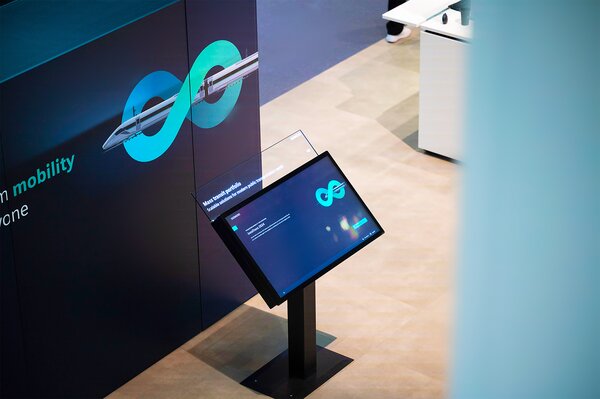 Interaktive Informationsstation auf einer Messe mit einem geneigten Touchscreen. Auf dem Bildschirm ist ein stilisiertes Unendlichkeitszeichen zu sehen, daneben steht Text zur nachhaltigen Mobilität. Im Hintergrund eine Messewand mit dem gleichen Symbol und dem Schriftzug „sustainable mobility for everyone“. Die Szene ist modern, reduziert und professionell beleuchtet.