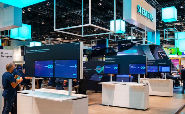  Großer Siemens-Messestand mit mehreren interaktiven Bildschirmen auf weißen Tischen, auf denen blaue Inhalte mit Unendlichkeitszeichen angezeigt werden. Im Hintergrund ist ein großes Siemens-Logo an der Decke beleuchtet, Besucher:innen stehen vor verschiedenen Stationen. Die Standarchitektur ist modern, mit offenen Rahmenstrukturen und blauen Leuchtwürfeln an der Decke.
