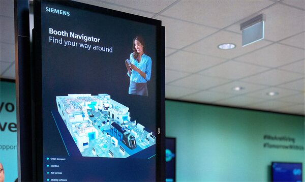 Digitaler Messestand-Navigator von Siemens auf einem großen Display. Die Visualisierung zeigt ein beleuchtetes 3D-Modell des Messestands mit verschiedenen Bereichen. Rechts oben ist eine Frau mit Tablet abgebildet, die das System bedient. Auf dem Display steht: „Booth Navigator – Find your way around“. Im Hintergrund ist eine hellgrüne Wand mit Hashtags und Deckenbeleuchtung sichtbar.