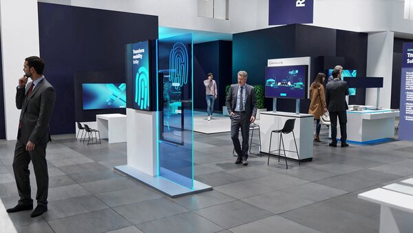 Offen gestalteter Messestand mit digitalen Touchscreens und interaktiven Stationen. Im Zentrum steht eine leuchtende Glasscheibe mit einem Fingerabdrucksymbol und dem Schriftzug „Transform security today“. Besucher:innen gehen umher oder betrachten die Exponate. Die Atmosphäre ist modern und geschäftlich, mit klaren Linien und dunklen Akzenten.