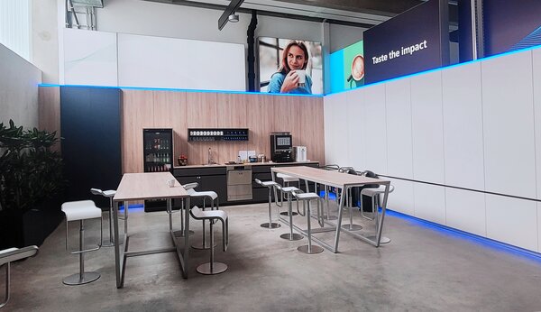 Heller Lounge-Bereich mit Kaffeemaschine, Tischen und Barhockern vor einer Holzrückwand; Bildschirm zeigt „Taste the impact“.