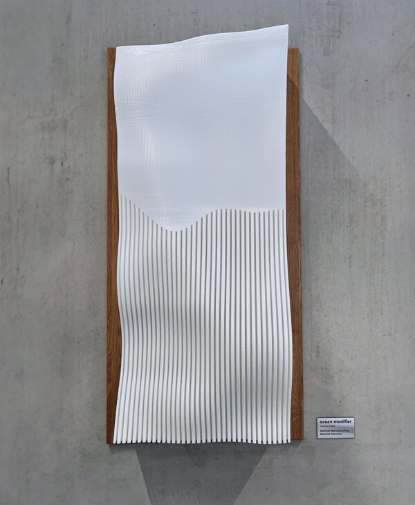 Weißes Wandrelief auf Holzplatte vor Betonwand, das mit Wellen und vertikalen Rillen ein abstrahiertes Meeresmotiv zeigt.