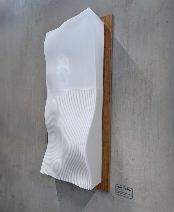 Weißes Wandrelief auf Holzplatte vor Betonwand, das mit Wellen und vertikalen Rillen ein abstrahiertes Meeresmotiv zeigt.