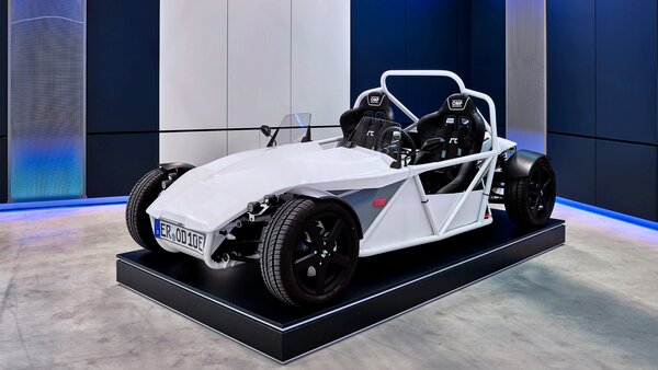 Weißer, offener Leichtbau-Sportwagen mit zwei Sitzen steht auf einer schwarzen Plattform in einem modernen Ausstellungsraum.
