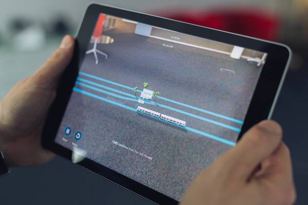Tablet zeigt eine AR-Visualisierung eines Zugs und Gebäudes auf einem Teppichboden mit blauen Linien.