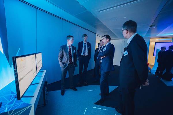 Männer in Anzügen stehen vor zwei Monitoren in einem blau ausgeleuchteten Ausstellungsraum.