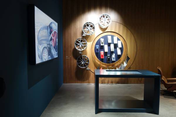 An einer gemütlichen Holzwand sind kreisförmig verschiedene Optionen für Felgen, Polster- und Außenfarben eines Autos präsentiert. Daneben stehen ein Tisch mit einem Touch-Screen und ein an der Wand hängender Monitor.