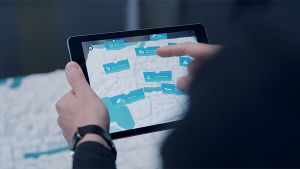 Tablet mit Übersichtskarte einer Stadt; die dargestellten Gebiete sind mit blauen Icons zu verschiedenen Themen (z.B. Car-Sharing, Verkehr, Umwelt) beschriftet.