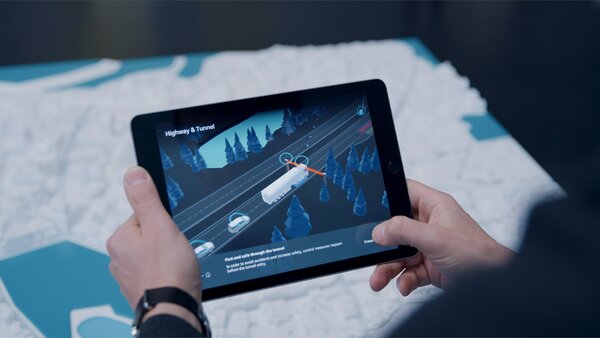 Person hält ein Tablet mit einer 3D-Visualisierung von Autobahn und Tunnel; das Display zeigt ein autonom fahrendes Fahrzeug in einer digitalen Verkehrssimulation.