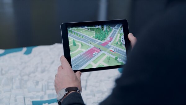 Tablet in den Händen eines Nutzers zeigt eine Kreuzung mit animierten Verkehrsflüssen in einer digitalen Stadtansicht.