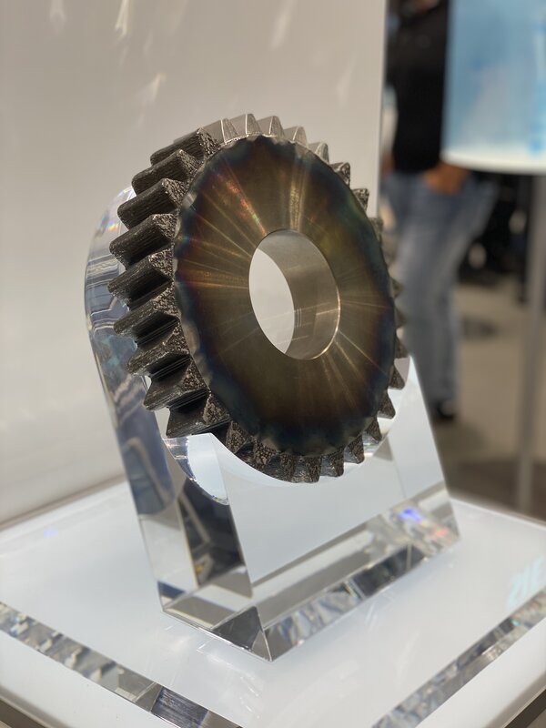 Metallisches Zahnrad mit bläulichen Anlassfarben steht auf einem transparenten Acrylständer in einer Ausstellung.